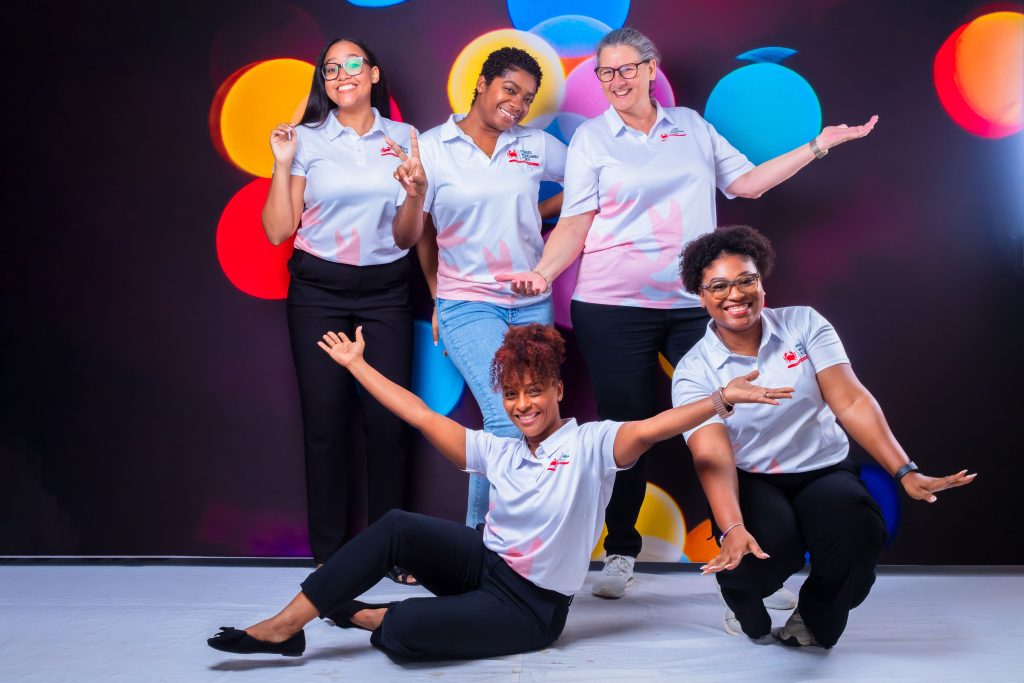 Five members of the Prinses Wilhelmina Fonds team in branded polo shirts, smiling and posing together in front of a backdrop of coloured lights
